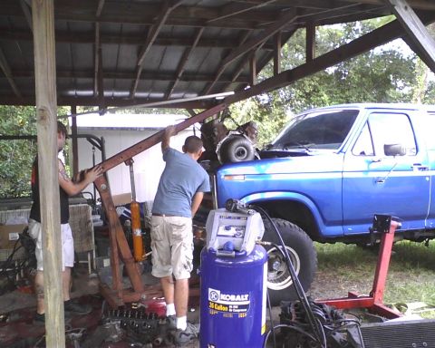1985 Ford Bronco - Mechanics work on a blue Ford Bronco under a covered structure. An engine hoist and air compressor are present.