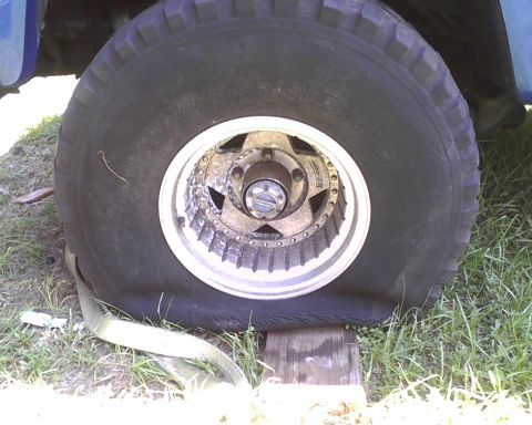 1985 Ford Bronco - Flat tire on a vehicle, supported by wood blocks and grass.