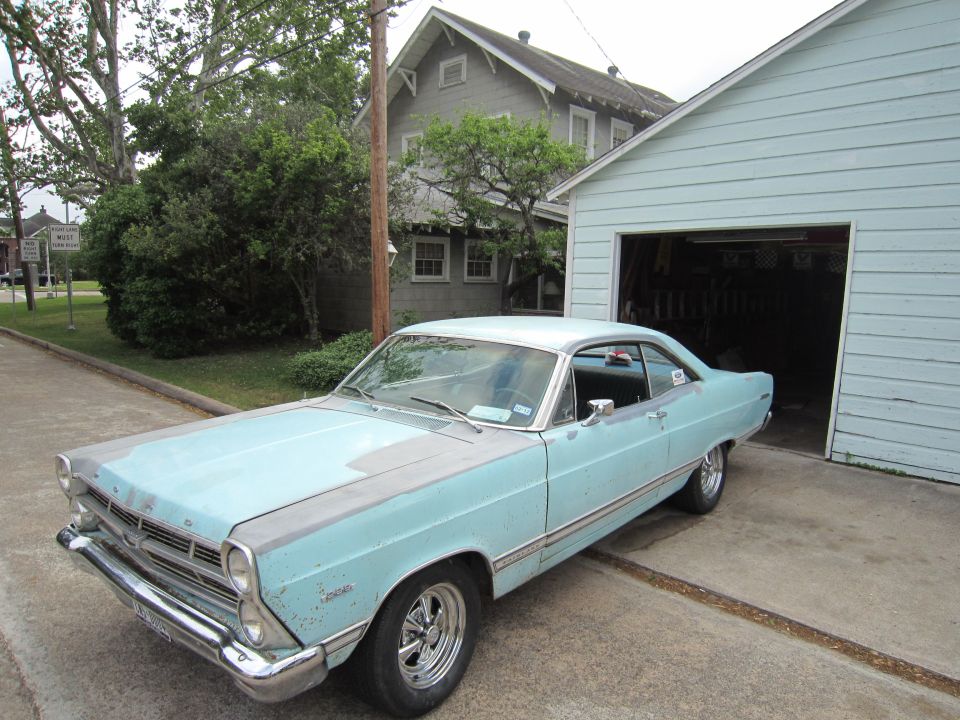 Light blue 1967 Ford Fairlane hardtop with primer spots parked by an open garage
