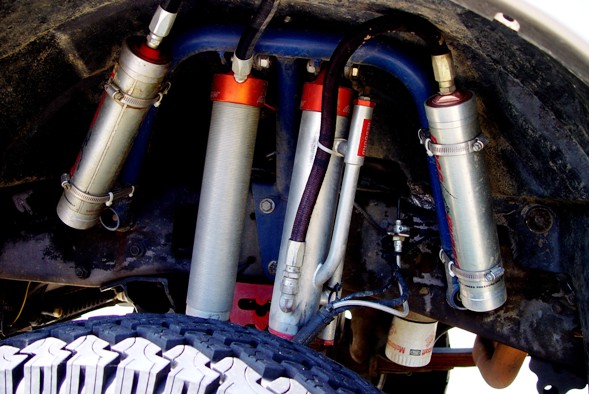 Underside view of dual coilover shocks per corner mounted in a wheel well above a mud tire