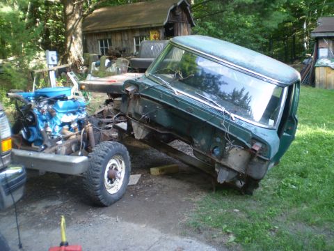 1979 Ford Bronco - A green truck cab and chassis, with a blue engine visible, sits in a yard with a wooden building in the background.