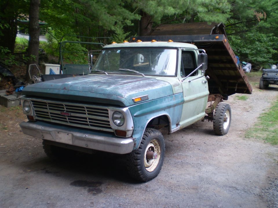1967 Ford dump truck with a raised bed, parked on a gravel driveway. Another vehicle is visible in the background