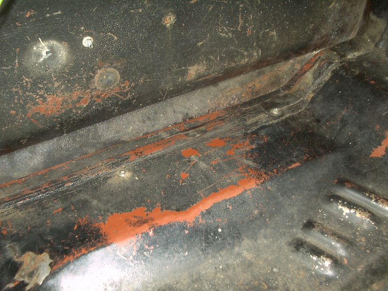 Rusty black floor pan with peeling paint exposing orange primer and rust.