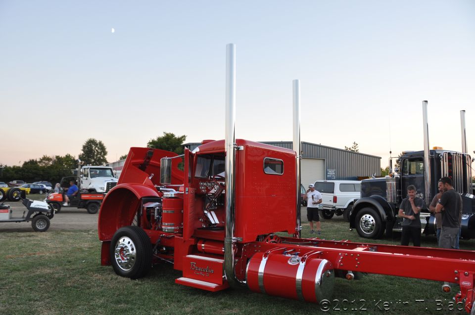 Red semi-truck chassis with large chrome exhaust stacks, hood open. Another black semi-truck is parked nearby