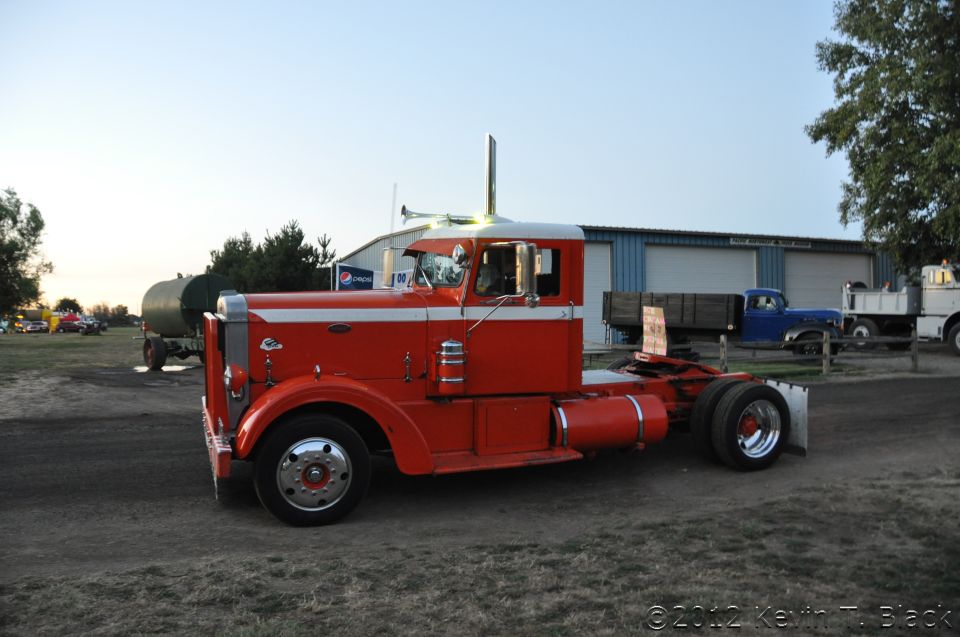 Red and white semi-truck, 3q front view. Other trucks and a building in background