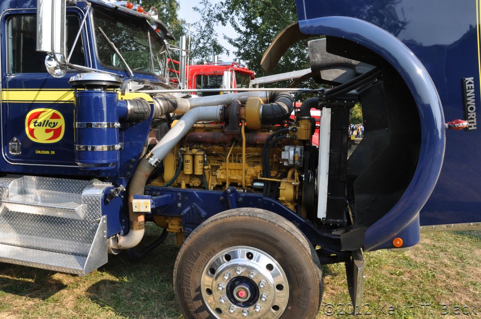 Blue Kenworth truck with hood open showing engine and front wheel. Another red truck is visible behind