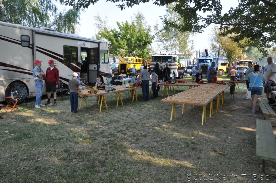 RV parked next to tables with toy trucks, with several large trucks in background