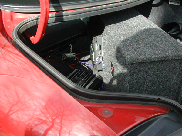Car trunk interior showing two audio amplifiers and a subwoofer box, with red exterior paint visible