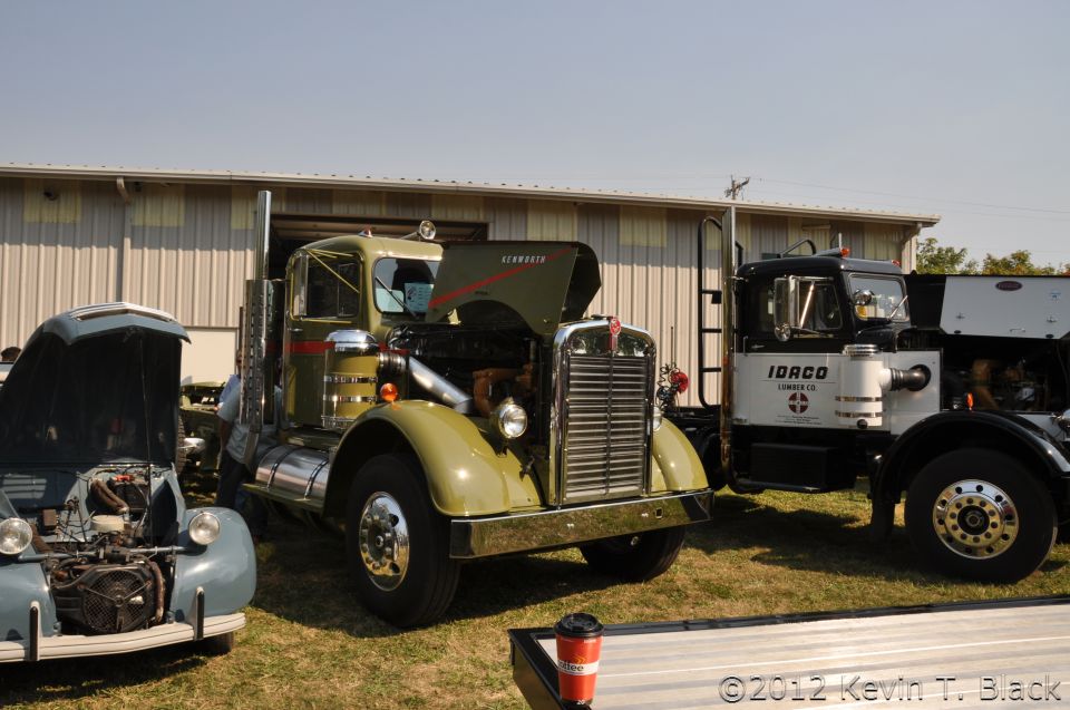 Green Kenworth truck front view, hood open, showing engine. Another truck to the right