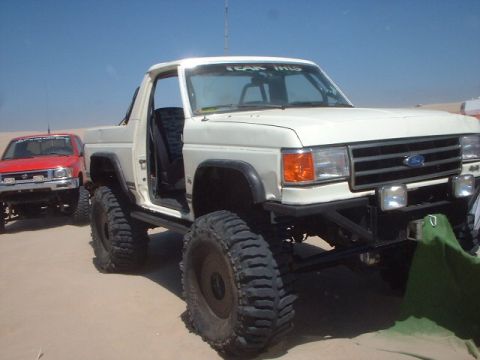 1991 Ford Bronco - Modified white Ford Bronco with large tires, seen with a red Toyota pickup in a sandy area.