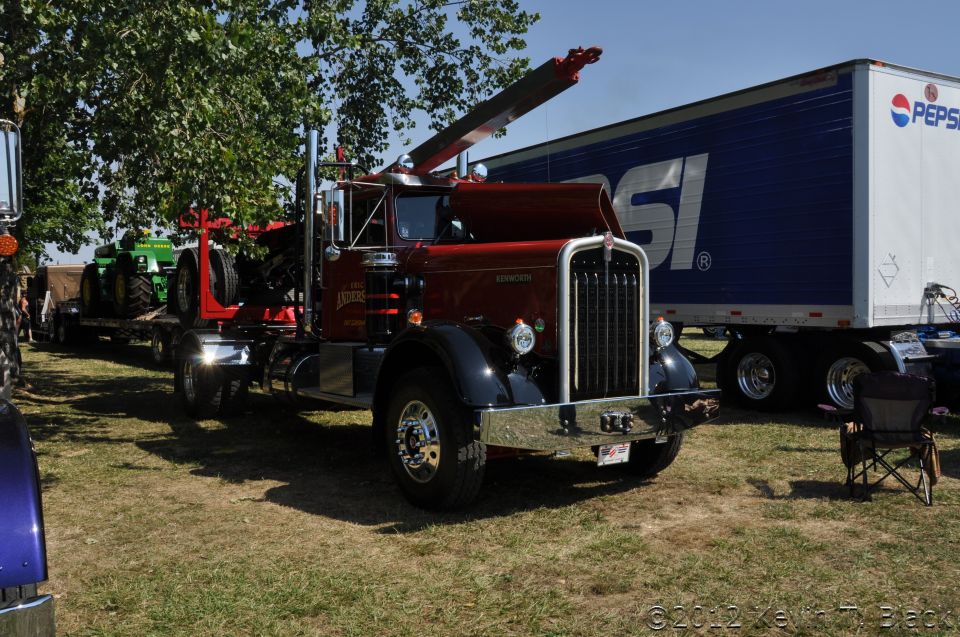 Red tow truck with a crane, parked next to a Pepsi trailer and a green tractor on a trailer