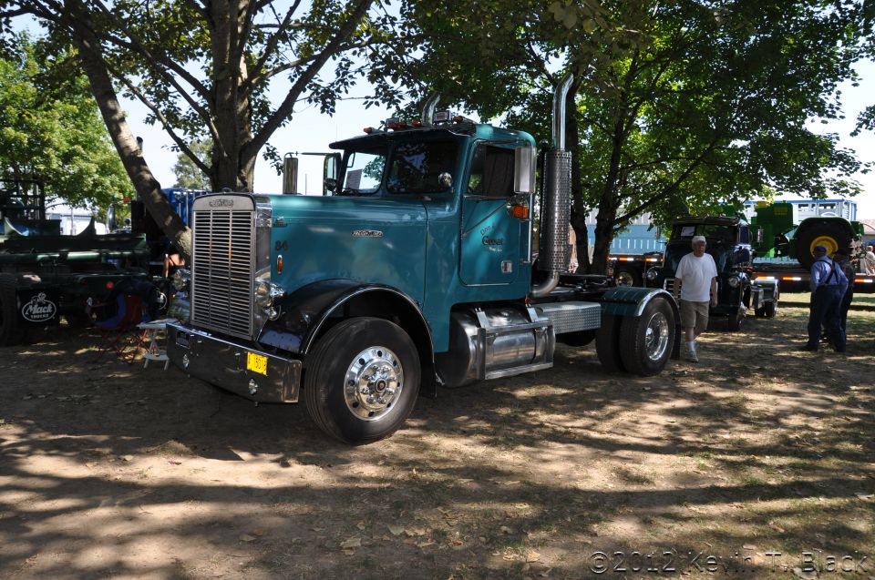 Teal semi-truck with chrome grille and stacks, parked outdoors under trees