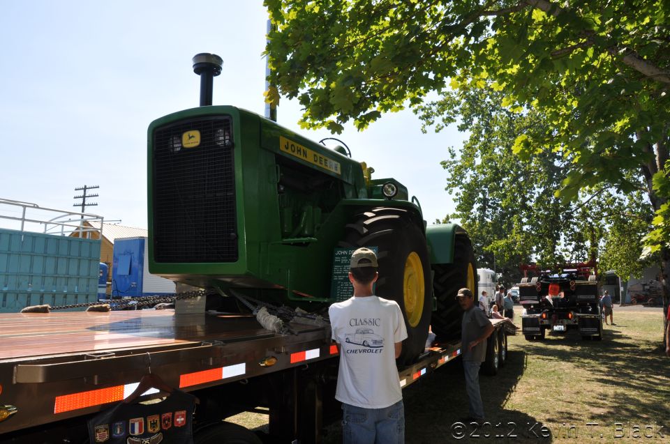 John Deere tractor being transported on a flatbed trailer, with another truck visible in the background