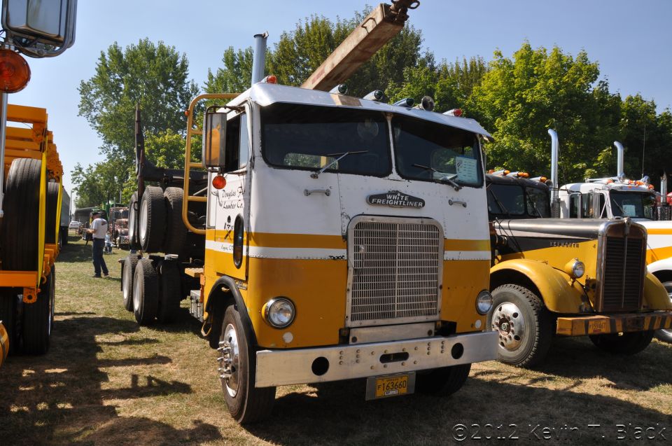 White Freightliner truck with yellow and white paint, crane boom extended. Another yellow truck is parked to its right