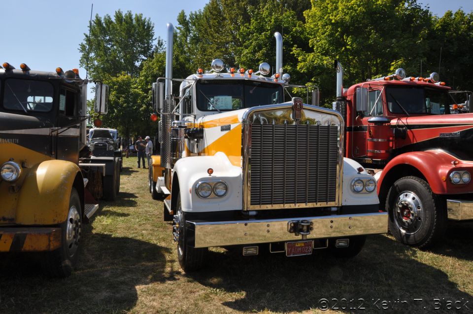 Front view of a white and yellow 1959 Kenworth W900A semi-truck, parked next to a red semi-truck and a yellow and black semi-truck