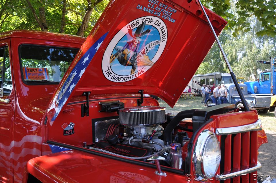 Red pickup truck with hood open showing engine and custom paint on the underside of the hood. Other vehicles visible in background