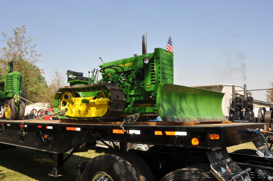 Green John Deere tractor with yellow tracks and a large green front loader on a black flatbed trailer