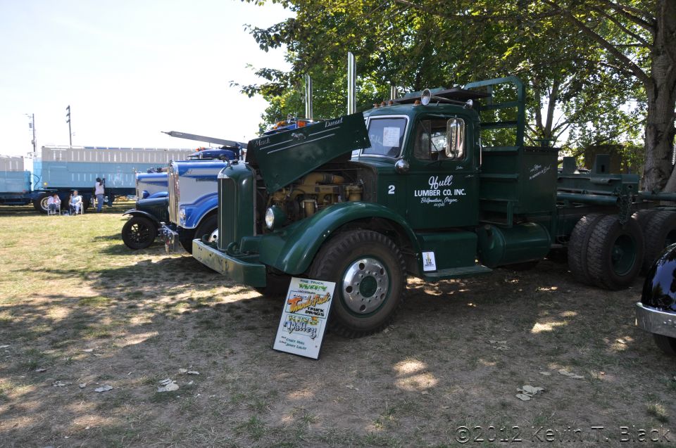 Green logging truck with hood open, "Hull Lumber Co. Inc." text on door, parked on grass next to a blue truck