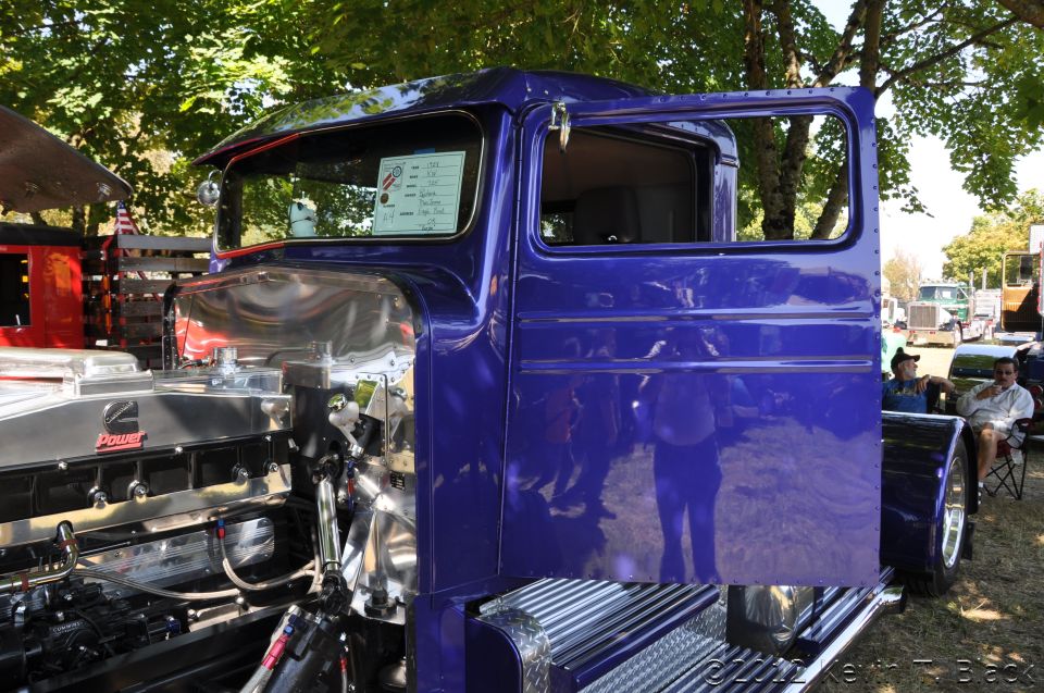 Purple semi-truck cab with open door, showing a large engine. Other trucks and people are visible in the background