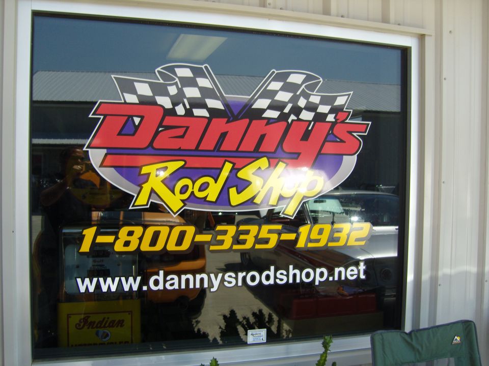 Window with "Danny's Rod Shop" logo, phone number, and website; reflection of a person and cars
