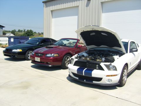 2007 Ford Mustang - Three Ford Mustangs, including a white model with its hood open, are parked in front of a garage.