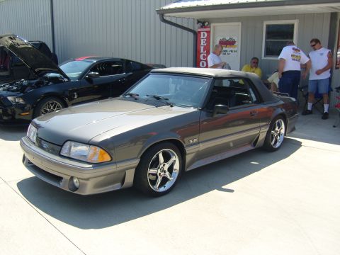 2007 Ford Mustang - A gray convertible Mustang is parked outside a building with a black Mustang in the background. People are standing near the entrance.