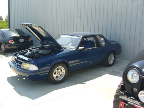 2007 Ford Mustang - A blue coupe with its hood open, parked next to other vehicles in front of a metal building.
