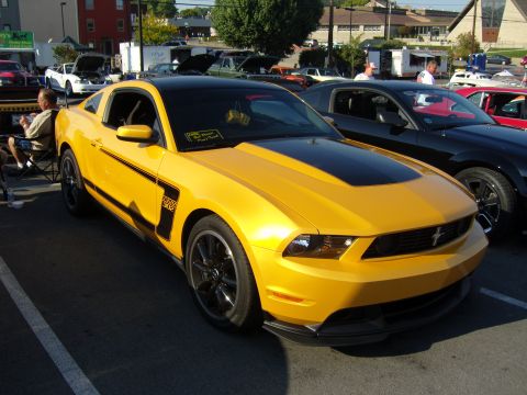 2007 Ford Mustang - yellow Ford Mustang Boss 302 with black side stripes parked at a car show among other cars and spectators.