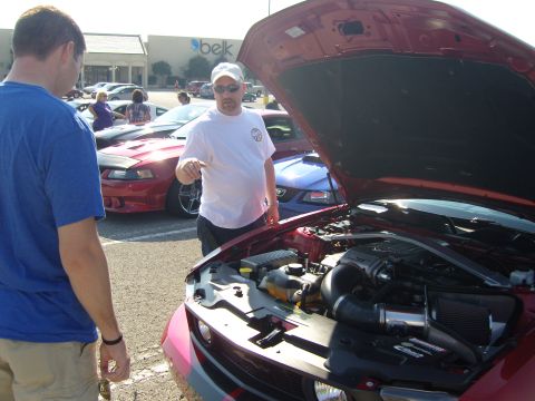 2007 Ford Mustang - Two men examine the engine bay of a red 2007 Ford Mustang at a car show.