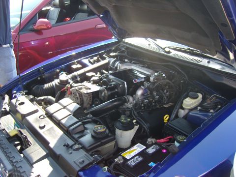 2007 Ford Mustang - Engine bay of a blue car, a supercharger and other engine components. A red car is visible in the background.