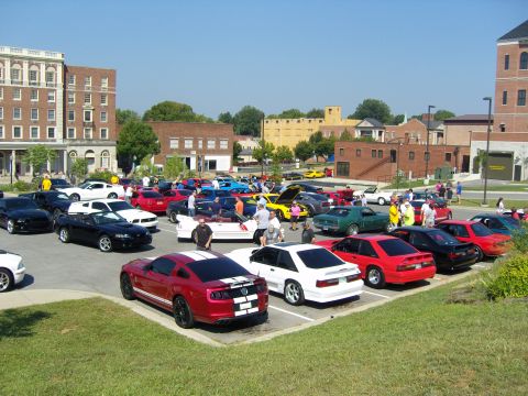 2007 Ford Mustang - Car show in a downtown lot filled with parked Mustangs and other cars, brick buildings in the background.