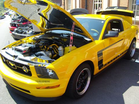 2007 Ford Mustang - A yellow Ford Mustang with its hood open, displaying the engine and custom details, parked at an outdoor car show.