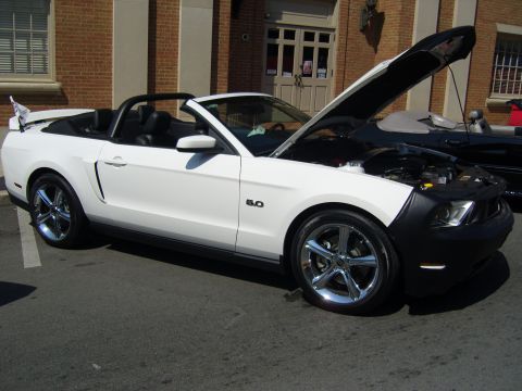 2007 Ford Mustang - A white convertible Mustang is parked outside a brick building with its hood open, revealing the engine bay.