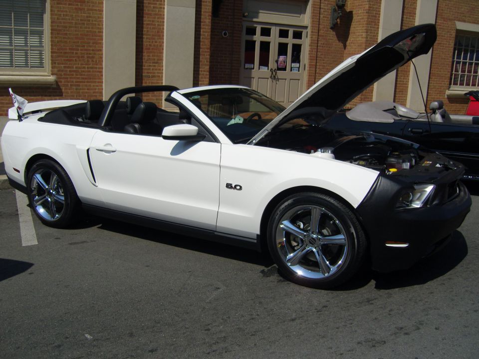 White convertible Mustang with the hood open, showing the engine. A black car is parked behind it