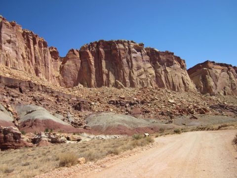 1991 Ford LTD Crown Victoria - A dirt road winds through a desert landscape with towering red rock cliffs under a clear blue sky.