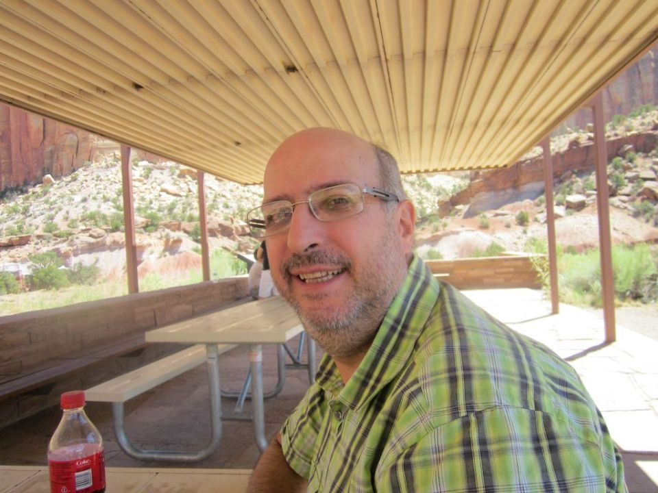 Man in green plaid shirt smiles under a covered picnic area with red rock formations in background