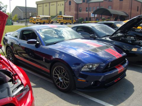 2007 Ford Mustang - A blue 2007 Ford Mustang with red racing stripes is parked at a car show, with other vehicles visible.