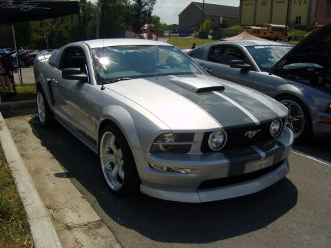 2007 Ford Mustang - A silver Ford Mustang with dual racing stripes is parked at an outdoor event.