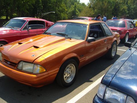 2007 Ford Mustang - An orange coupe with aftermarket hood scoops and racing wheels is parked in a lot with other vehicles.
