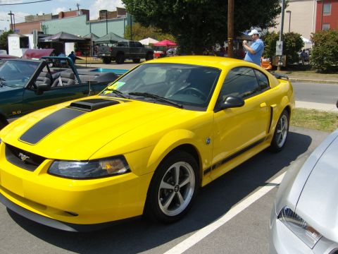 2007 Ford Mustang - A yellow Ford Mustang with black racing stripes is parked at an outdoor car show, with other vehicles and tents in the background.