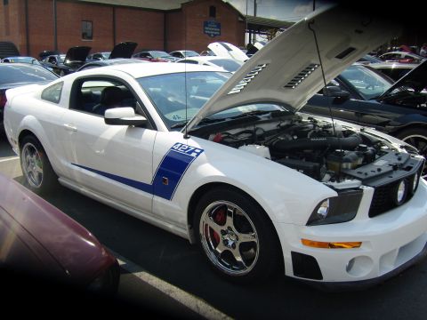 2007 Ford Mustang - A white Ford Mustang with blue racing stripes is parked with its hood open at a car show.