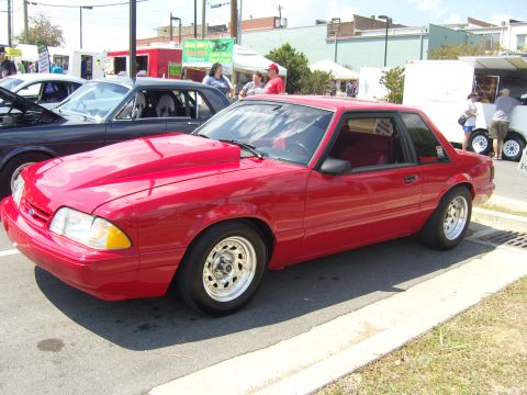 2007 Ford Mustang - A red coupe with a prominent hood scoop and aftermarket wheels is parked at an outdoor event.