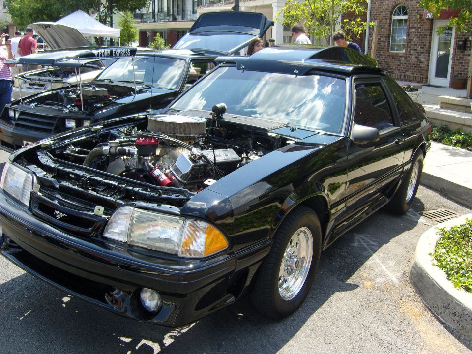 Black Ford Mustang from the late 80s with hood open showing engine. Another Mustang is behind it