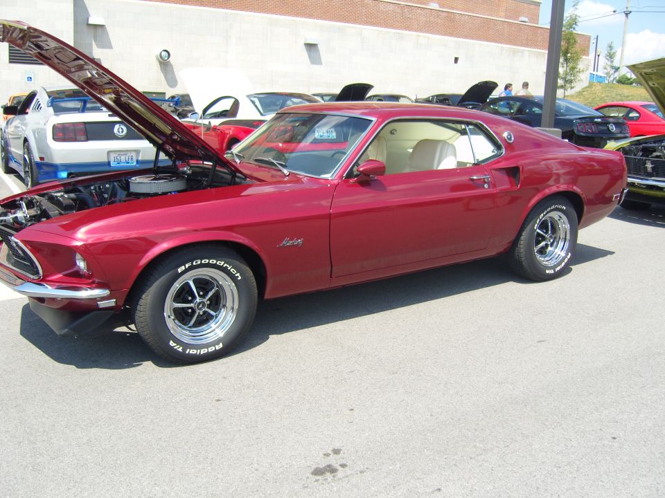 Red 1969 Ford Mustang with hood open, parked at a car show. Other cars are visible in the background