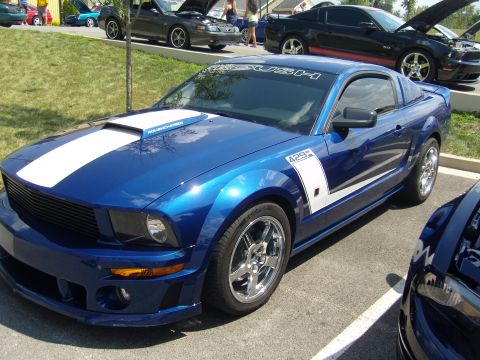 2007 Ford Mustang - A blue Ford Mustang with racing stripes is parked at an outdoor car show. Other Mustangs are visible in the background.