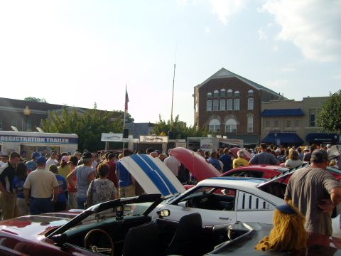 2007 Ford Mustang - A crowd mills around vehicles at an outdoor event, with a registration trailer in the background under a bright sky.