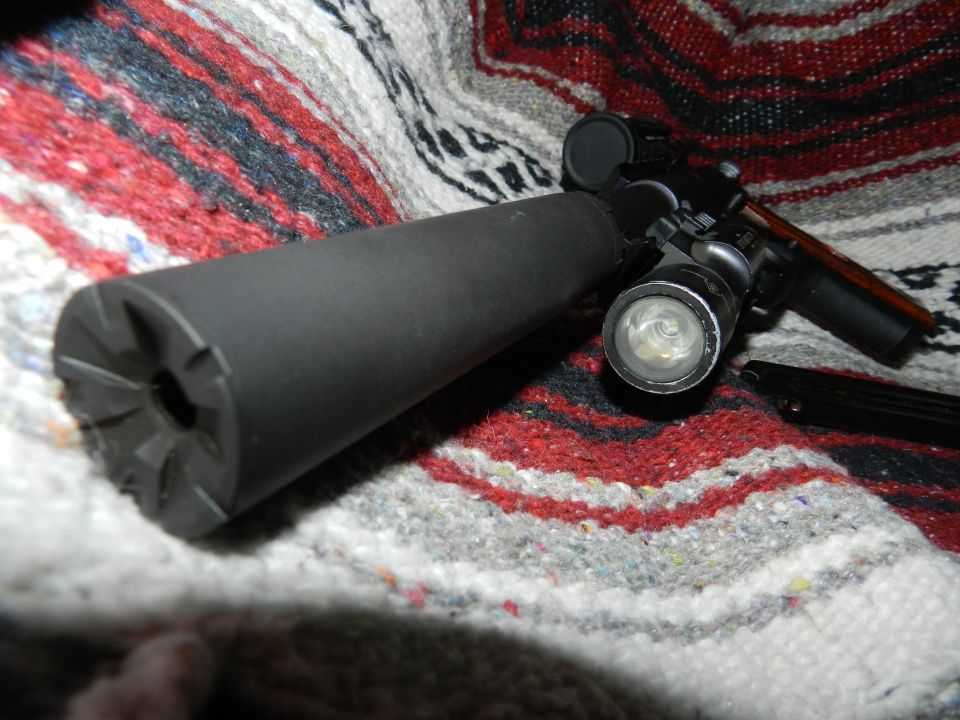 Firearm with a suppressor attached, a flashlight mounted above it, and a magazine to the side, resting on a patterned blanket