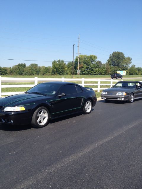 2007 Ford Mustang - Two Ford Mustangs, a black coupe and a gray convertible, are parked on an asphalt surface with a white fence and trees in the background.