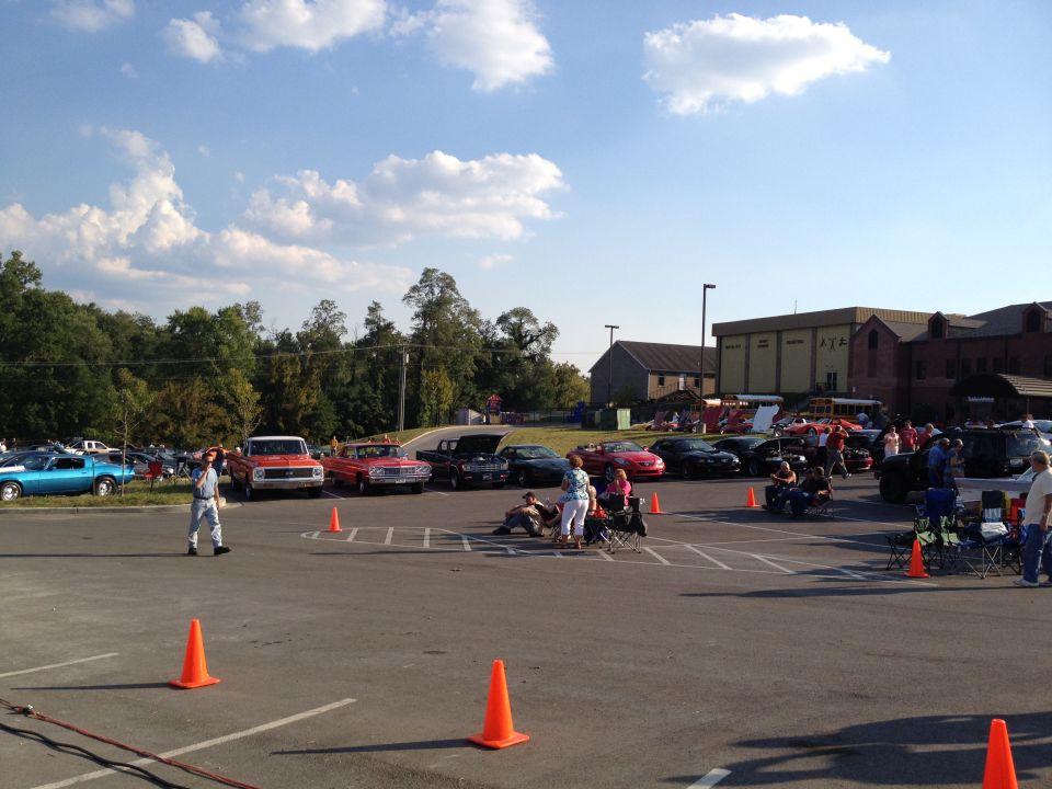 Parking lot scene with various classic cars and people gathered under a blue sky