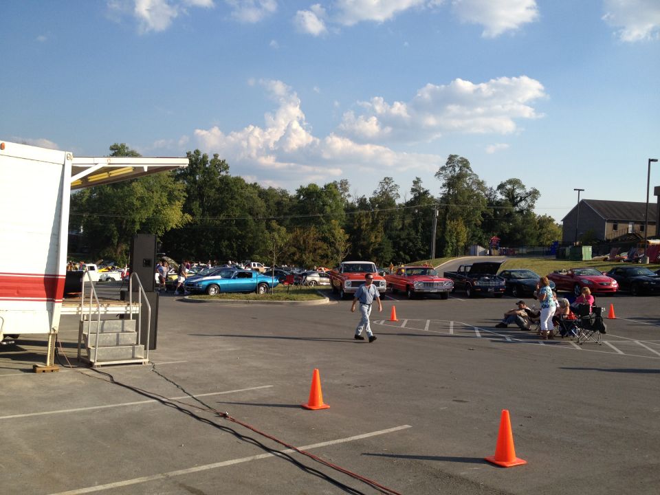 Outdoor car show with a stage on the left and many cars parked in rows under a blue sky with clouds